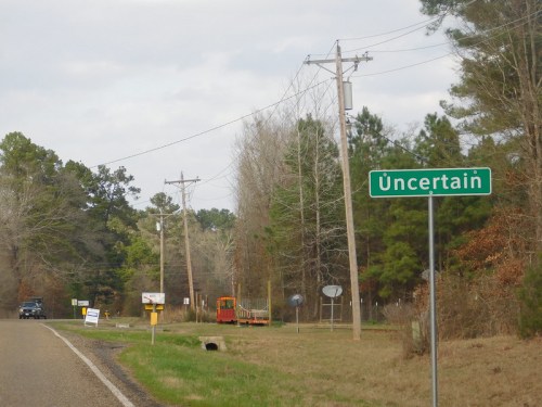 A roadside sign reads "Uncertain," with trees and power lines in the background along a rural road.