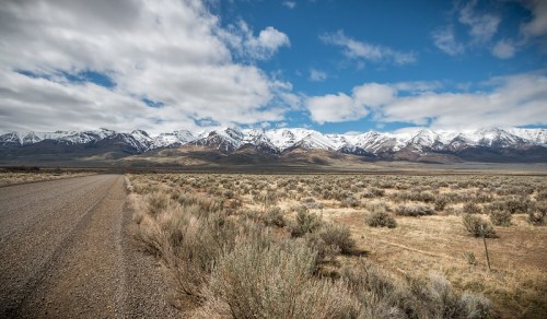 A gravel road leads through a valley with snow-capped mountains under a partly cloudy sky. Sagebrush covers the foreground.