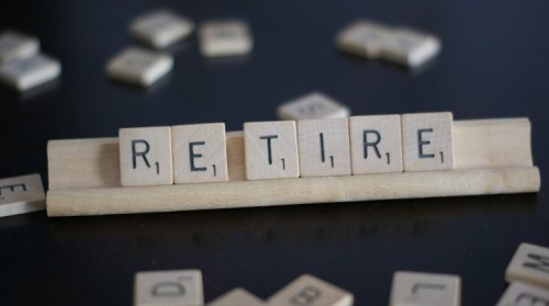 Scrabble tiles spell out the word "RETIRE" on a wooden rack, with scattered tiles in the background.