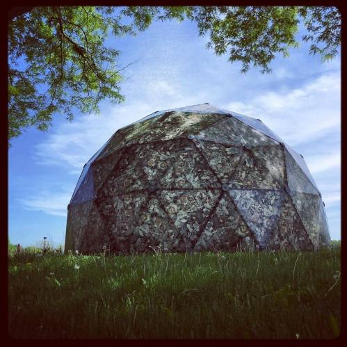 A geodesic dome structure covered in natural materials, set against a blue sky and green grass.