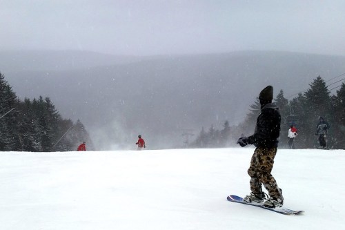 A snowy ski slope with snowboarders and skiers in winter gear, surrounded by trees and a misty mountain backdrop.