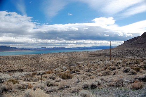 A vast landscape featuring dry terrain, sparse vegetation, and a lake under a cloudy sky. Power lines are visible in the distance.