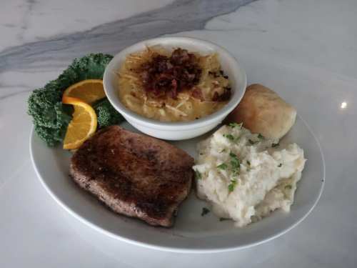 A plate with a grilled pork chop, creamy macaroni and cheese, mashed potatoes, a biscuit, kale, and an orange slice.