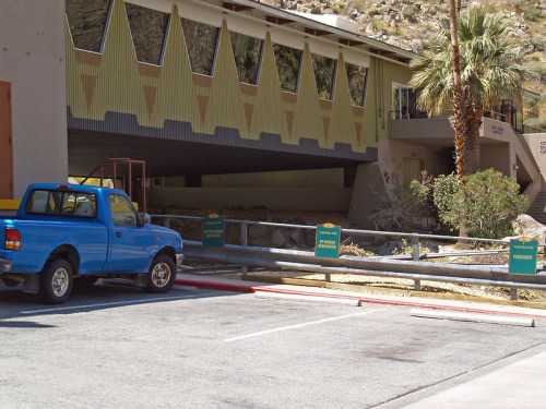 A blue pickup truck parked near a building with a unique architectural design and signs indicating parking regulations.