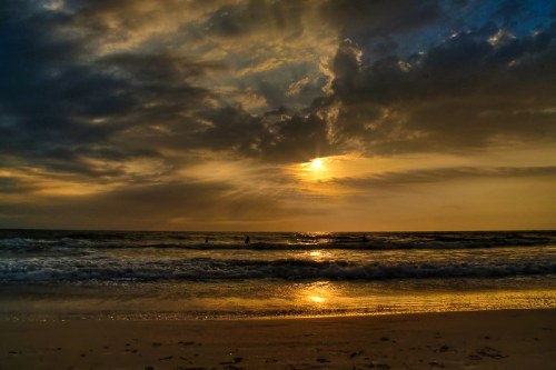 Sunset over the ocean with dramatic clouds and gentle waves reflecting golden light on the shore.