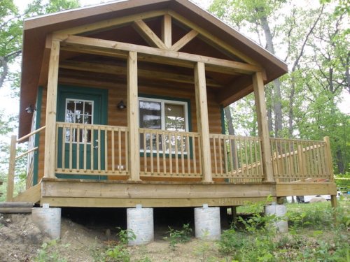 A wooden cabin with a porch, surrounded by trees, set on stilts in a natural landscape.