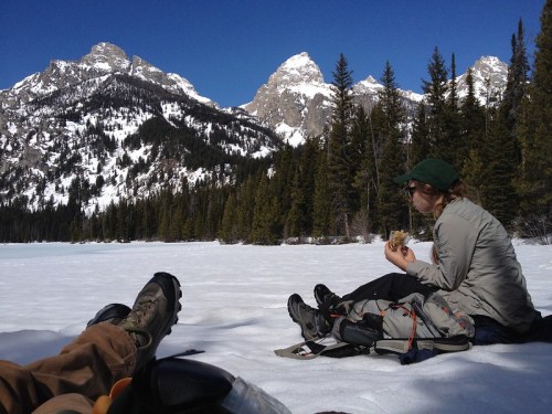 A person sits on snow by a lake, eating, with mountains and pine trees in the background under a clear blue sky.