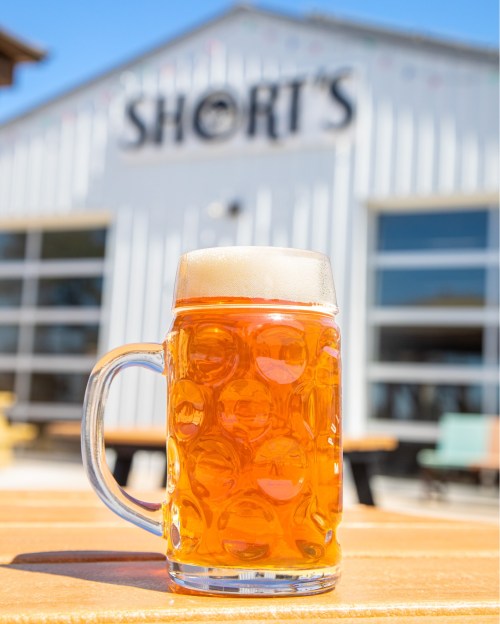 A frosty mug of amber beer sits on a table, with a brewery building in the background.