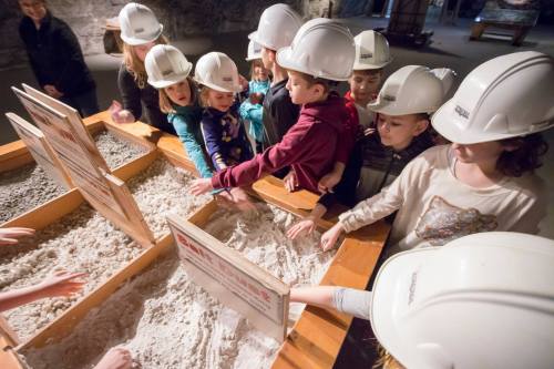Children wearing hard hats explore sand tables with informational signs in a museum setting.
