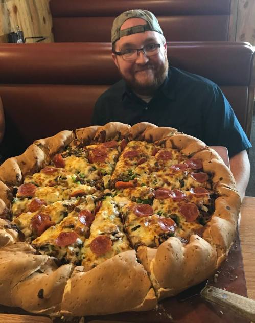 A man smiles beside a large, stuffed crust pizza topped with pepperoni and various vegetables.