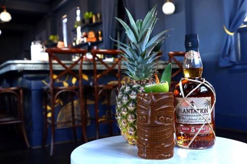 A tiki mug and a bottle of rum beside a pineapple on a table, with a bar in the background.