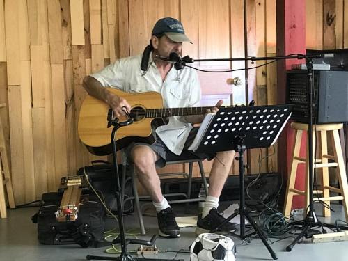 A man plays acoustic guitar and sings into a microphone, seated with music sheets in a wooden room.