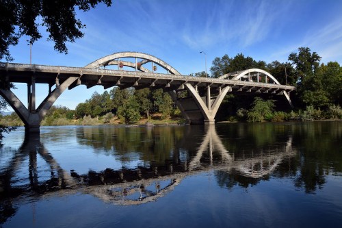 A concrete bridge arches over a calm river, reflecting its structure in the water beneath a clear blue sky.