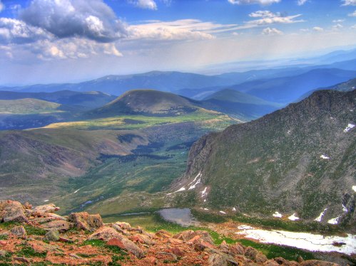 A panoramic view of rolling mountains and valleys under a partly cloudy sky, with patches of snow and greenery.