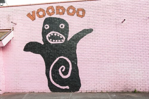 A large black voodoo doll mural with a spiral on its belly, painted on a pink brick wall. The word "VOODOO" is above it.