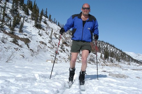 A man skiing in shorts on a snowy landscape, wearing a blue jacket and ski poles, with trees in the background.