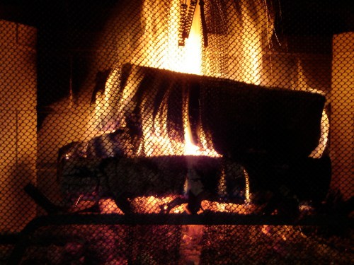 A cozy fireplace with logs burning brightly behind a metal grate, casting warm light and shadows.