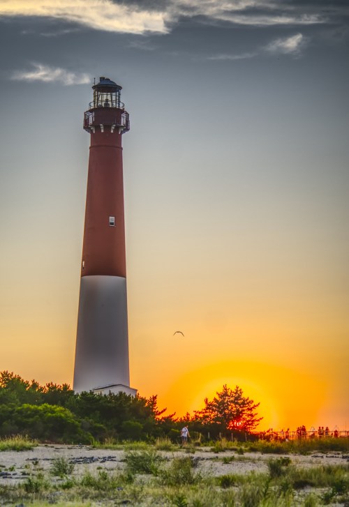A tall lighthouse stands against a vibrant sunset, surrounded by greenery and a sandy beach.