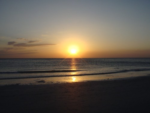 A serene beach at sunset, with the sun reflecting on calm waters and a clear sky.