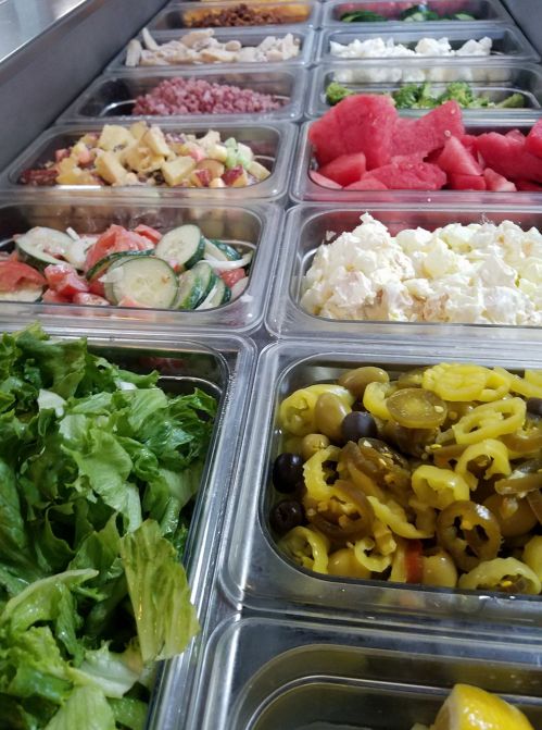 A variety of fresh salad ingredients in containers, including greens, vegetables, fruits, and toppings.