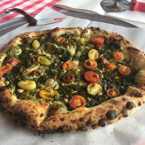 A freshly baked pizza topped with pesto, cherry tomatoes, and pine nuts on a rustic table with a checkered cloth.