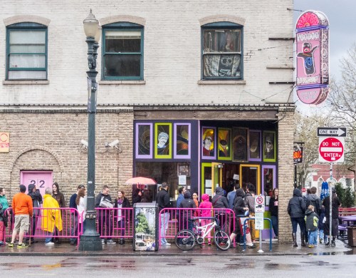 A colorful donut shop with a long line of people waiting outside on a rainy day. Bright signage and umbrellas are visible.