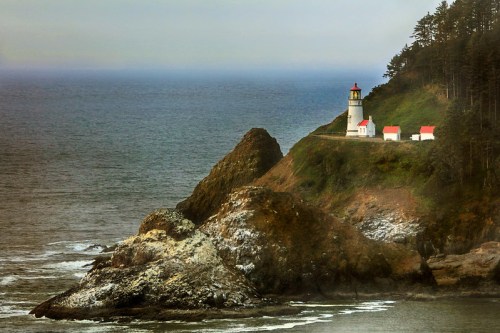 A lighthouse perched on a rocky cliff overlooking the ocean, surrounded by trees and a misty atmosphere.