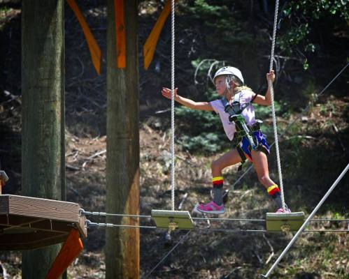 A young girl in a helmet balances on a rope bridge, surrounded by trees and wooden structures.