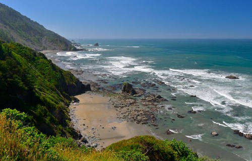 A scenic coastal view with rocky shores, waves crashing, and lush green cliffs under a clear blue sky.