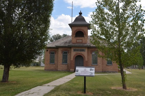 Historic brick building with a tower, surrounded by trees and grass, featuring an informational sign in front.