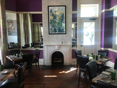 Cozy restaurant interior with purple walls, wooden tables, black chairs, and a colorful artwork above a fireplace.
