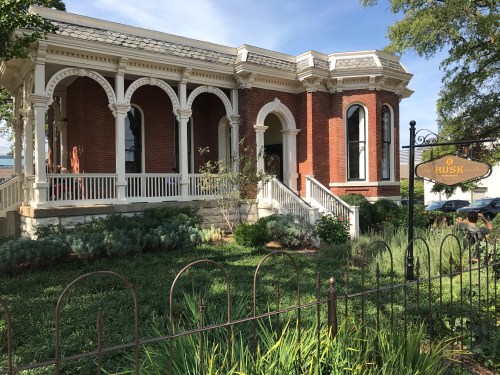 Historic brick building with arched windows, a porch, and a sign reading "Husk," surrounded by greenery and a fence.
