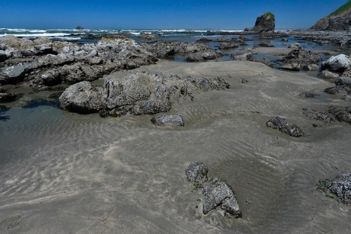 Rocky shoreline with sandy patches, gentle waves, and a clear blue sky in the background.