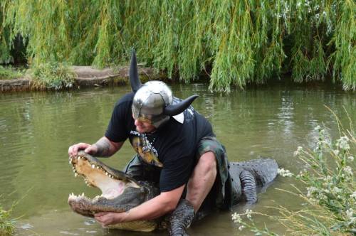 A person in a Viking helmet sits on a crocodile, holding its jaws open near a pond surrounded by greenery.