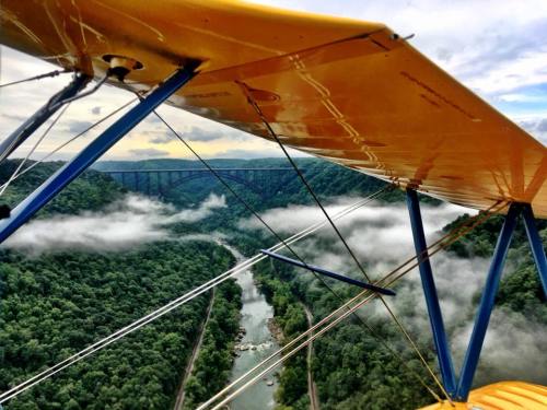 View from a biplane wing over a lush green valley with a river and a bridge, surrounded by misty clouds.