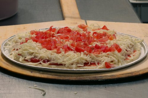 A pizza topped with shredded cheese and diced tomatoes on a metal tray, resting on a wooden pizza peel.