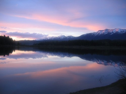 A serene lake reflects a colorful sunset with mountains in the background and trees lining the shore.