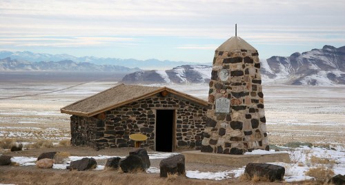 A stone monument beside a small building in a vast, snowy landscape with mountains in the background.
