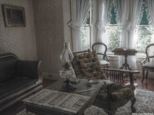 A vintage living room with a patterned armchair, an oil lamp, and large windows draped with lace curtains.