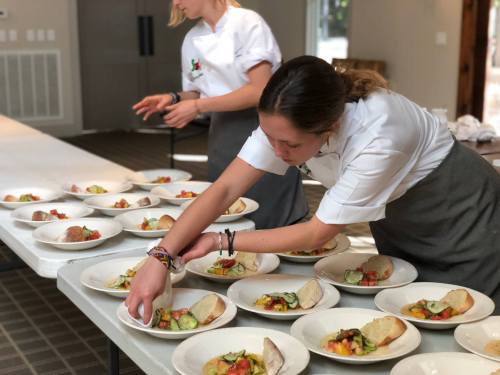 Two chefs plating dishes with vegetables and bread on a long table in a bright kitchen setting.