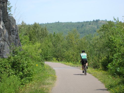 A cyclist rides along a paved path surrounded by lush greenery and rocky terrain under a clear blue sky.