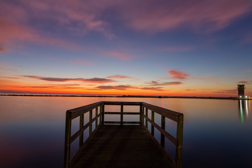 A wooden pier extends into calm waters at sunset, with vibrant colors in the sky and a distant tower illuminated.