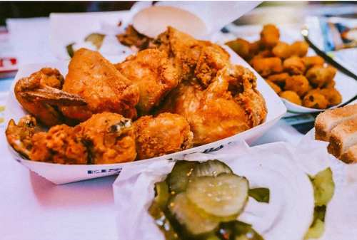 A platter of crispy fried chicken with pickles, bread, and a side of fried snacks on a table.