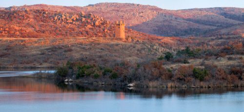 A scenic view of a lake surrounded by rocky hills and autumn-colored trees, featuring a stone tower on the shore.