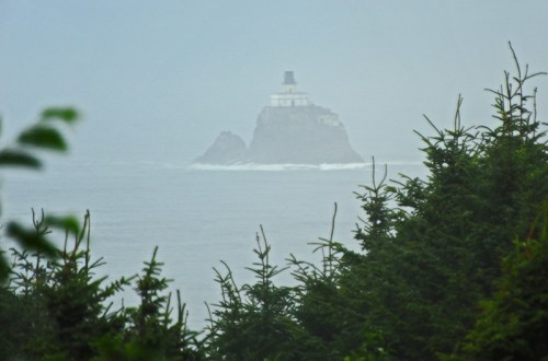 A foggy view of a lighthouse on a rocky island, surrounded by ocean and framed by green trees in the foreground.