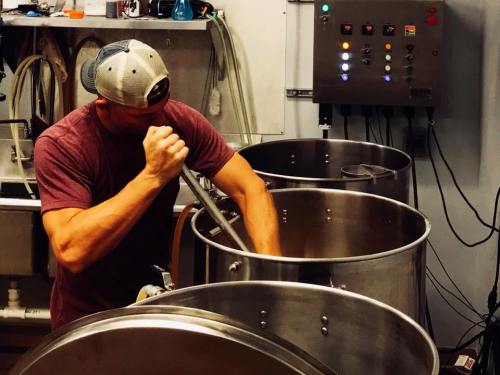 A person stirring a large metal brewing kettle in a brewery, with equipment and controls in the background.