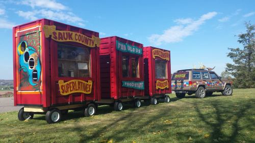 Colorful food stands on wheels beside a decorated vehicle, set against a clear blue sky and green grass.