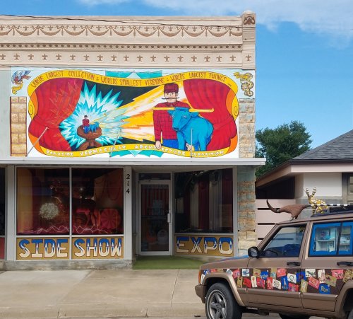 Colorful storefront featuring a sideshow theme with murals, showcasing unique attractions and a decorated vehicle outside.
