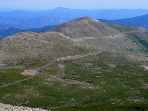 A winding road leads up a grassy mountain, with distant peaks visible under a clear blue sky.