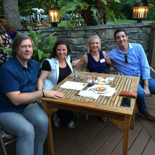 A group of four people sitting at a wooden table outdoors, enjoying drinks and appetizers in a lush garden setting.